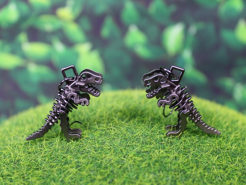 May include: Two black metal dinosaur skeleton charms, facing each other, on a green grassy surface.