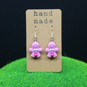 K&ouml;nnte beinhalten: Rosa Lebkuchenmann-Ohrringe mit einer wei&szlig;en Schleife und rosa Kn&ouml;pfen. Die Ohrringe befinden sich auf einer braunen Karte mit dem Text "hand made".