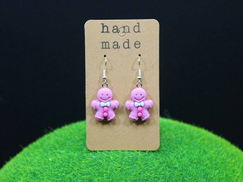 K&ouml;nnte beinhalten: Rosa Lebkuchenmann-Ohrringe mit einer wei&szlig;en Schleife und rosa Kn&ouml;pfen. Die Ohrringe befinden sich auf einer braunen Karte mit dem Text "hand made".