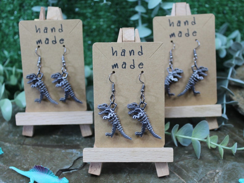 May include: Three pairs of silver dinosaur skeleton earrings on brown card stock with the text "hand made" on each card.
