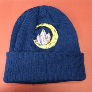 May include: A navy blue knitted beanie with a yellow crescent moon and crystal design embroidered on the front.