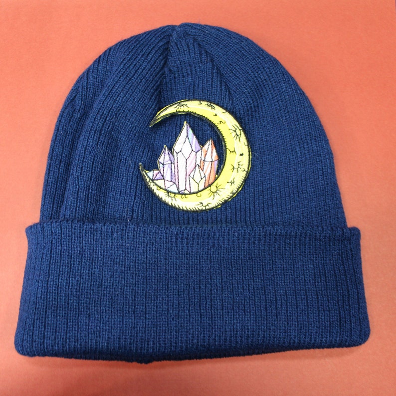 May include: A navy blue knitted beanie with a yellow crescent moon and crystal design embroidered on the front.