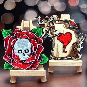 May include: Two embroidered iron-on patches. One patch features a red rose with a white skull in the centre. The other patch features a stylised ace of hearts playing card with flames.