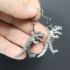 May include: A pair of silver dinosaur skeleton earrings. The earrings are made of metal and feature a detailed dinosaur skeleton design.