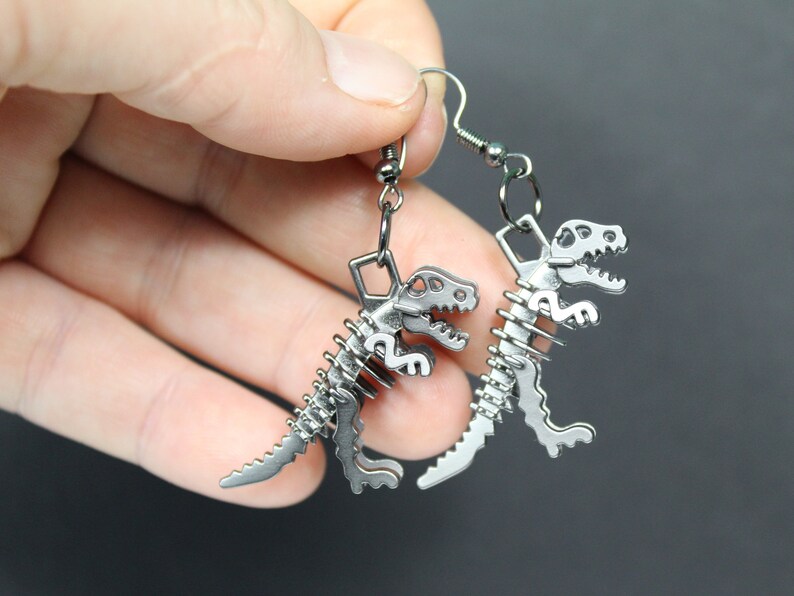 May include: A pair of silver dinosaur skeleton earrings. The earrings are made of metal and feature a detailed dinosaur skeleton design.