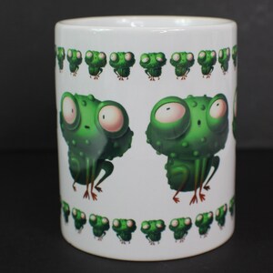 May include: White ceramic mug featuring a green frog design. The frog has large eyes and a surprised expression. The mug is decorated with a row of smaller frogs around the top and bottom.