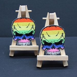 May include: Two rainbow skull patches displayed on miniature wooden easels. The patches feature a skull design with a rainbow-striped top, blue eye sockets, and a pink mouth. Set against a dark backdrop.