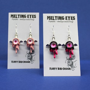 May include: Two pairs of novelty earrings with a melting eye design. The earrings are pink and black with silver hooks. The text "MELTING EYES" and "Flyballs - Always watching!" is printed on the backing card.