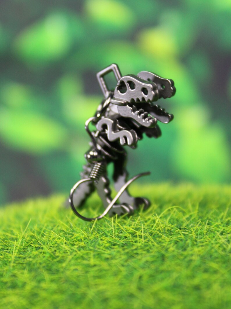 May include: Silver dinosaur skeleton earrings with a springy wire hook fastening. The earrings are on a green grassy surface.