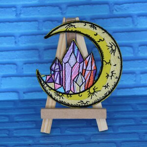 May include: A yellow crescent moon with a black outline and a sun symbol inside. The moon is filled with purple, pink, and orange crystals. The patch is displayed on a small wooden easel.