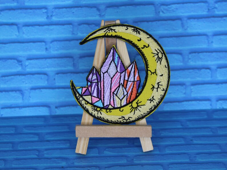 May include: A yellow crescent moon with a black outline and a sun symbol inside. The moon is filled with purple, pink, and orange crystals. The patch is displayed on a small wooden easel.