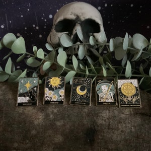 May include: Five gold-toned enamel pins depicting tarot cards. The cards are The Star, The Sun, The Moon, Death, and The Wheel of Fortune. The pins are set against a dark wood background.