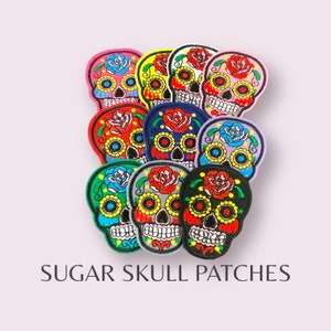 May include: A set of ten colourful embroidered sugar skull patches. The patches are in various colours, including pink, yellow, blue, green, red, and purple. Each patch features a sugar skull with a floral design and intricate details.