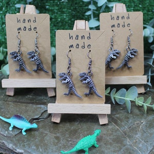 May include: Three pairs of silver dinosaur skeleton earrings displayed on small wooden easels. Each easel has a brown card with the text "hand made" on it.