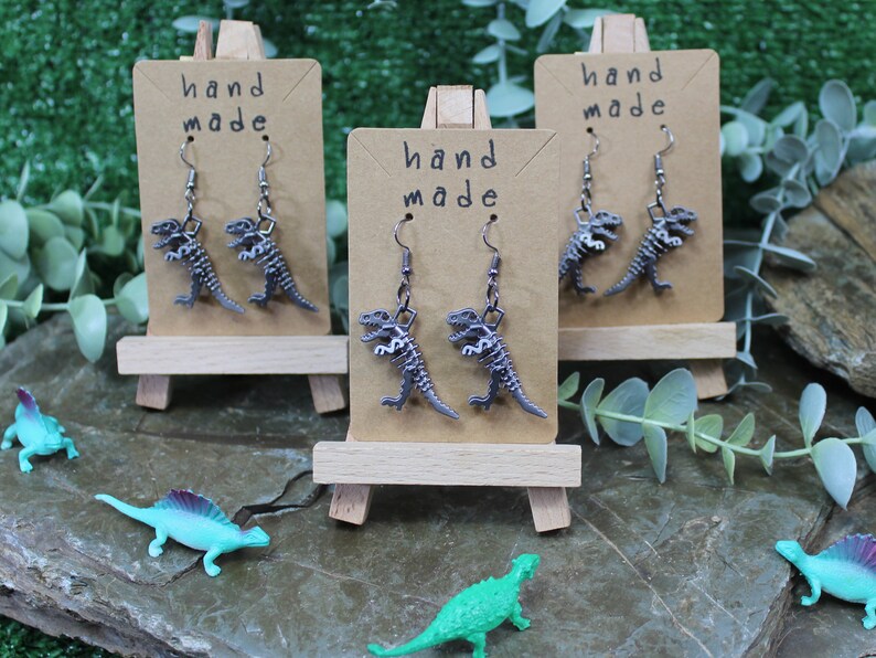 May include: Three pairs of silver dinosaur skeleton earrings displayed on small wooden easels. Each easel has a brown card with the text "hand made" on it.