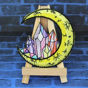 May include: A yellow crescent moon with a sun and stars design, embroidered with a cluster of colorful crystals in the center.