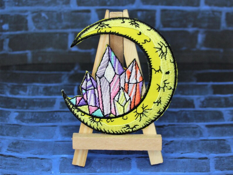 May include: A yellow crescent moon with a sun and stars design, embroidered with a cluster of colorful crystals in the center.
