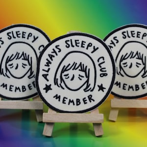May include: Three circular patches with the text "ALWAYS SLEEPY CLUB MEMBER" in black on a white background. Each patch features a cartoon illustration of a sleepy face. The patches are displayed on small wooden easels against a rainbow background.