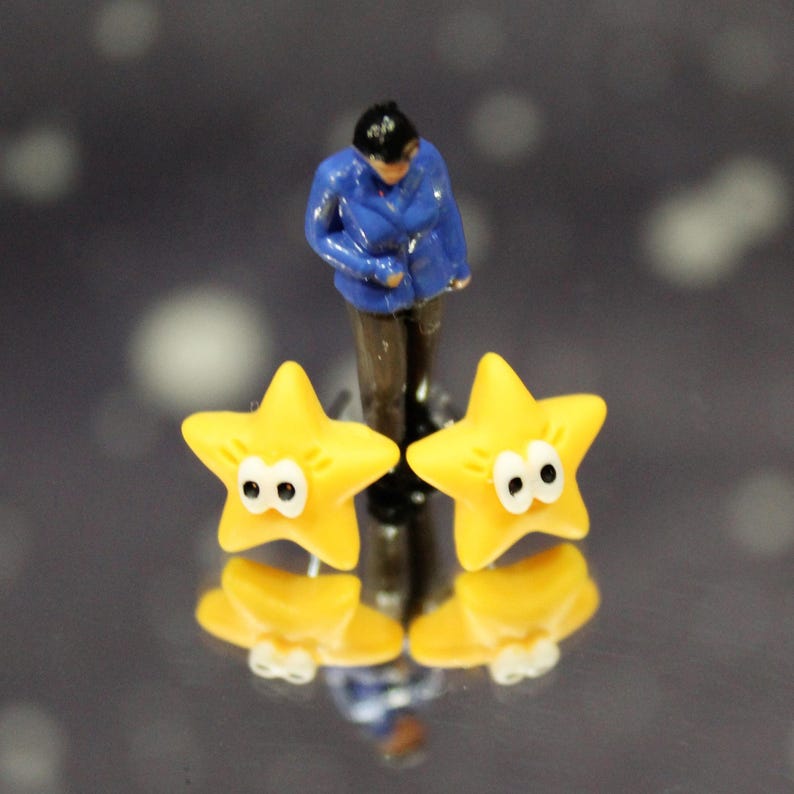 Single yellow star earring with googly eyes displayed outdoors on moss, with a small figure for scale.• Style: yellow star stud earrings
• Design: star stud earrings, petite and lightweight look
• Color: sunny yellow
• Occasion: everyday wear, gifts, party, festival, stocking stuffer