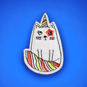 May include: White embroidered cat with a rainbow tail and a unicorn horn. The cat has a red eye patch and a pink nose.