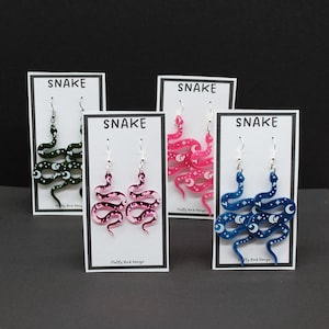 May include: Four pairs of dangle earrings featuring a snake design with white stars and moons. The earrings are displayed on white cards with the word "SNAKE" printed on them. The snakes are in green, pink, and blue.