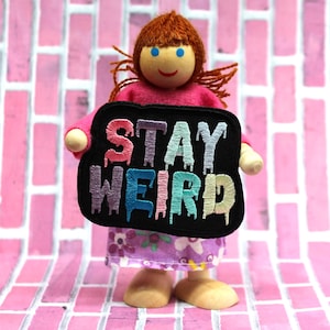 May include: A wooden doll with red hair and blue eyes holds a black patch with the words "STAY WEIRD" embroidered in rainbow colours.