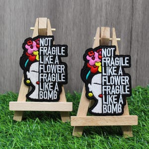 May include: Two embroidered patches with a Frida Kahlo-inspired design. The patches feature a woman's profile with a floral headdress and the text "NOT FRAGILE LIKE A FLOWER FRAGILE LIKE A BOMB" in white on a black background. They are displayed on small wooden easels.