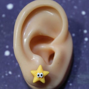 A yellow cartoon-style star earring with googly eyes, worn on a mannequin ear. Small, lightweight, and playful design.