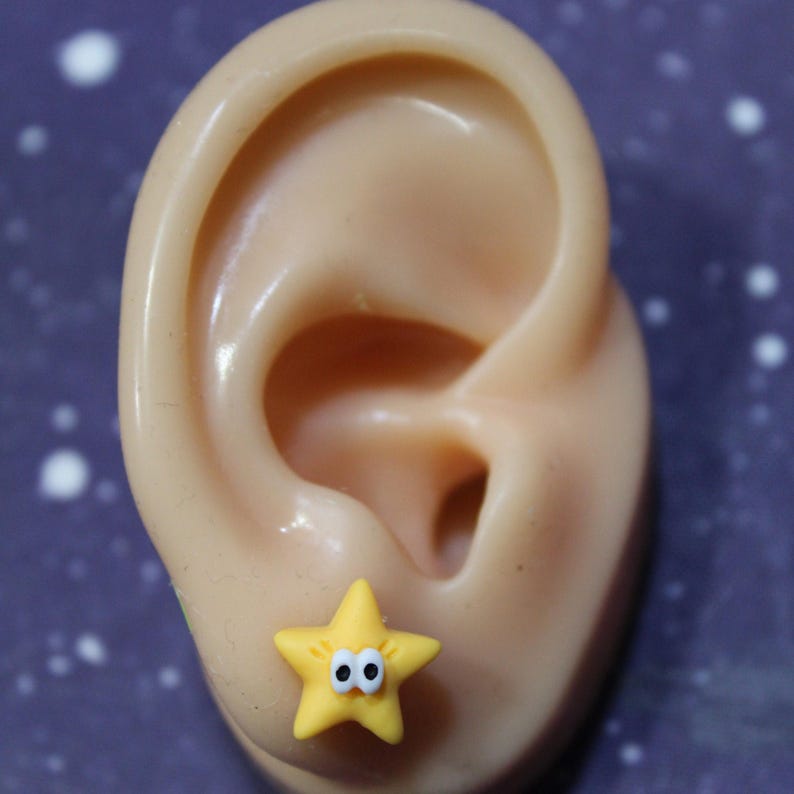A yellow cartoon-style star earring with googly eyes, worn on a mannequin ear. Small, lightweight, and playful design.