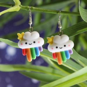 May include: A pair of cloud-shaped earrings with rainbow detailing. Each earring has a white cloud with a smiling face, a yellow star, and a rainbow. The earrings are suspended from silver-coloured hooks.
