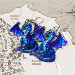 May include: A pair of blue and purple dragon earrings. The dragons are made of clear acrylic and have a stained glass look. The earrings are hanging from silver hooks.