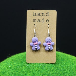 K&ouml;nnte beinhalten: Lilafarbene Lebkuchenmann-Ohrringe mit rosa Schleifen und wei&szlig;en Kn&ouml;pfen. Die Ohrringe befinden sich auf einer braunen Karte mit dem Text "hand made".