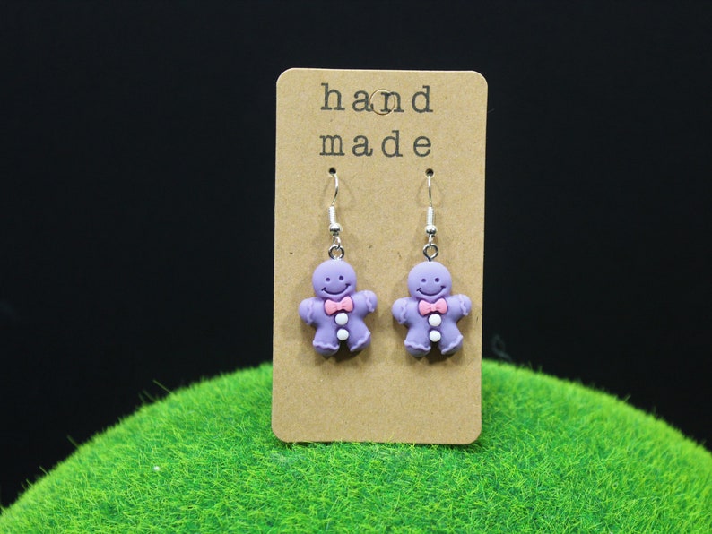 K&ouml;nnte beinhalten: Lilafarbene Lebkuchenmann-Ohrringe mit rosa Schleifen und wei&szlig;en Kn&ouml;pfen. Die Ohrringe befinden sich auf einer braunen Karte mit dem Text "hand made".