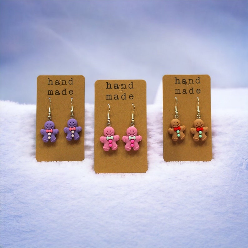 K&ouml;nnte beinhalten: Drei Paar handgemachte Lebkuchenmann-Ohrringe. Die Ohrringe befinden sich auf braunen Karten mit dem Text "hand made". Die Lebkuchenm&auml;nner sind lila, rosa und braun.