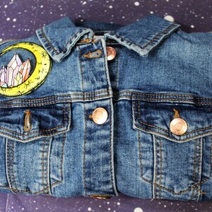 May include: A blue denim jacket with a yellow crescent moon and crystal patch on the left side. The jacket has two front pockets and a button closure.