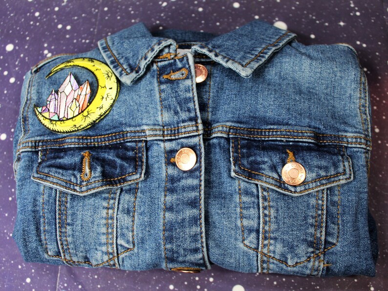 May include: A blue denim jacket with a yellow crescent moon and crystal patch on the left side. The jacket has two front pockets and a button closure.