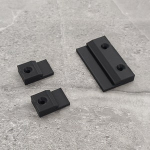 Puede incluir: Tres soportes de estantería de plástico negro con agujeros para tornillos. El soporte más grande es rectangular y tiene un borde en un lado. Los dos soportes más pequeños son cuadrados.