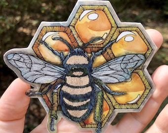 May include: A sticker featuring a detailed illustration of a bee with blue and tan stripes, set against a honeycomb background. The bee has detailed wings and legs. The sticker is held in a hand, with a blurred natural background.