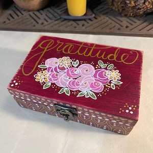 May include: A burgundy wooden box with the word "gratitude" in gold script. The box is decorated with pink roses and floral accents. The sides feature a gold and white pattern, and it has a metal clasp.