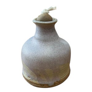 May include: A ceramic oil lamp with a rounded body and a narrow neck. The upper portion features a speckled light purple glaze, while the lower section displays a smooth, pale yellow finish. A small, round, tan stopper with a white wick is inserted.