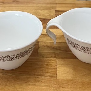 Puede incluir: Dos cuencos de sopa de cerámica blanca con un diseño floral marrón alrededor de la base y un asa curva. Los cuencos están sobre una superficie de madera.