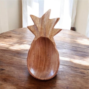 May include: A wooden pineapple-shaped bowl with a natural wood grain finish. The bowl has a deep, oval-shaped interior and a starburst-like top, resembling a pineapple crown. The bowl is likely used for serving or display.