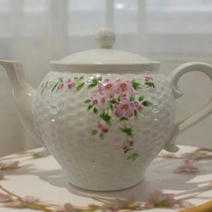 May include: A white ceramic teapot featuring a textured basket-weave pattern on the body and a delicate hand-painted floral design with pink blossoms and green leaves. The teapot includes a matching lid with a small round knob handle.