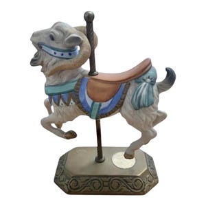 May include: A carousel ram figurine with a cream-colored body, brown saddle, and decorative blue and green accents. The base is bronze-colored with an ornate design, and a central pole supports the ram.