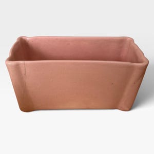 May include: A rectangular ceramic planter in a solid dusty pink hue. The vessel features a smooth, matte finish with slightly flared corners and a visible vertical hairline crack extending down the left side of the front exterior wall.