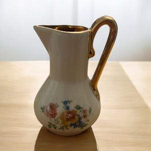 May include: Cream-colored ceramic pitcher with a gold handle and rim. The pitcher features a floral design with pink, yellow, and blue flowers. The handle curves gracefully from the top to the base of the pitcher.