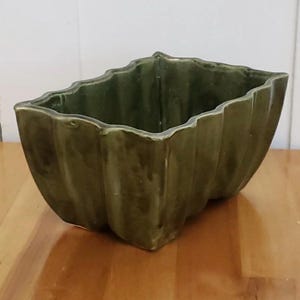 May include: A rectangular ceramic planter with a glossy olive green glaze and a scalloped, wavy rim. The exterior features vertical fluted detailing along the sides, creating a textured appearance with subtle variations in the green color tone.