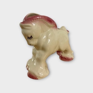 May include: A vintage ceramic horse figurine in a cream color with pink accents on the mane and hooves. The horse has closed eyes and a small opening on its back, suggesting it may be a planter or decorative piece.