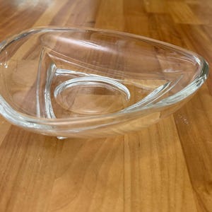 Op de afbeelding: Transparante glazen serveerschaal met een uniek, asymmetrisch ontwerp. De schaal is verdeeld in drie secties, met een centraal cirkelvormig gebied en twee gebogen compartimenten. Het glas is transparant, waardoor de inhoud zichtbaar is. De schaal staat op een houten oppervlak.