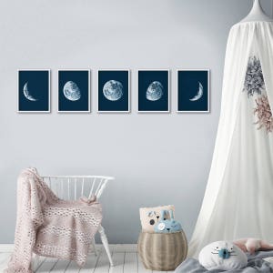 May include: Five framed prints of the moon in different phases, displayed on a light blue wall. The prints are in a dark blue color scheme and feature a white border.